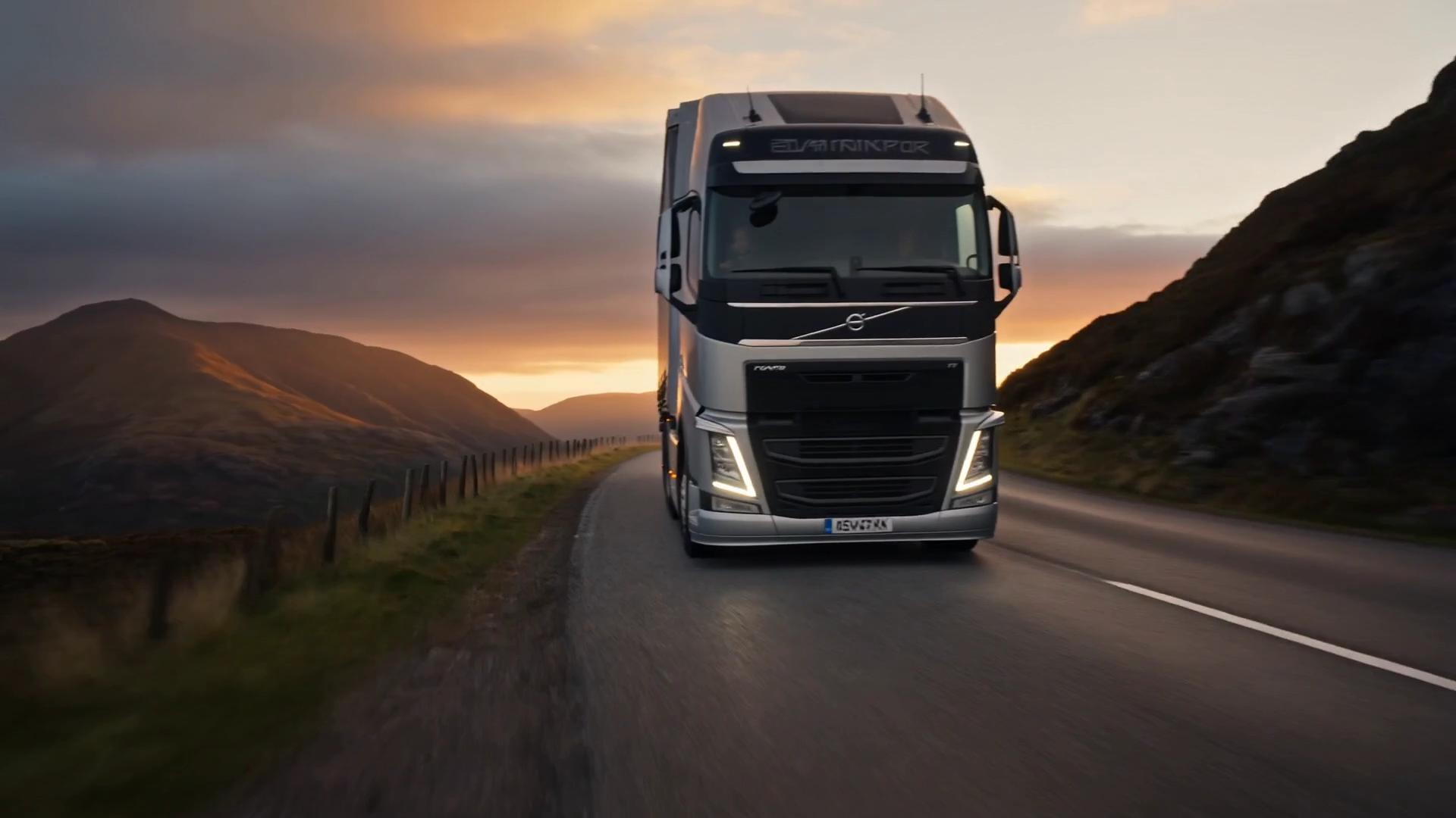 A truck on a Highland road at dusk, headlights lit, mountains silhouetted against a fading amber sky.