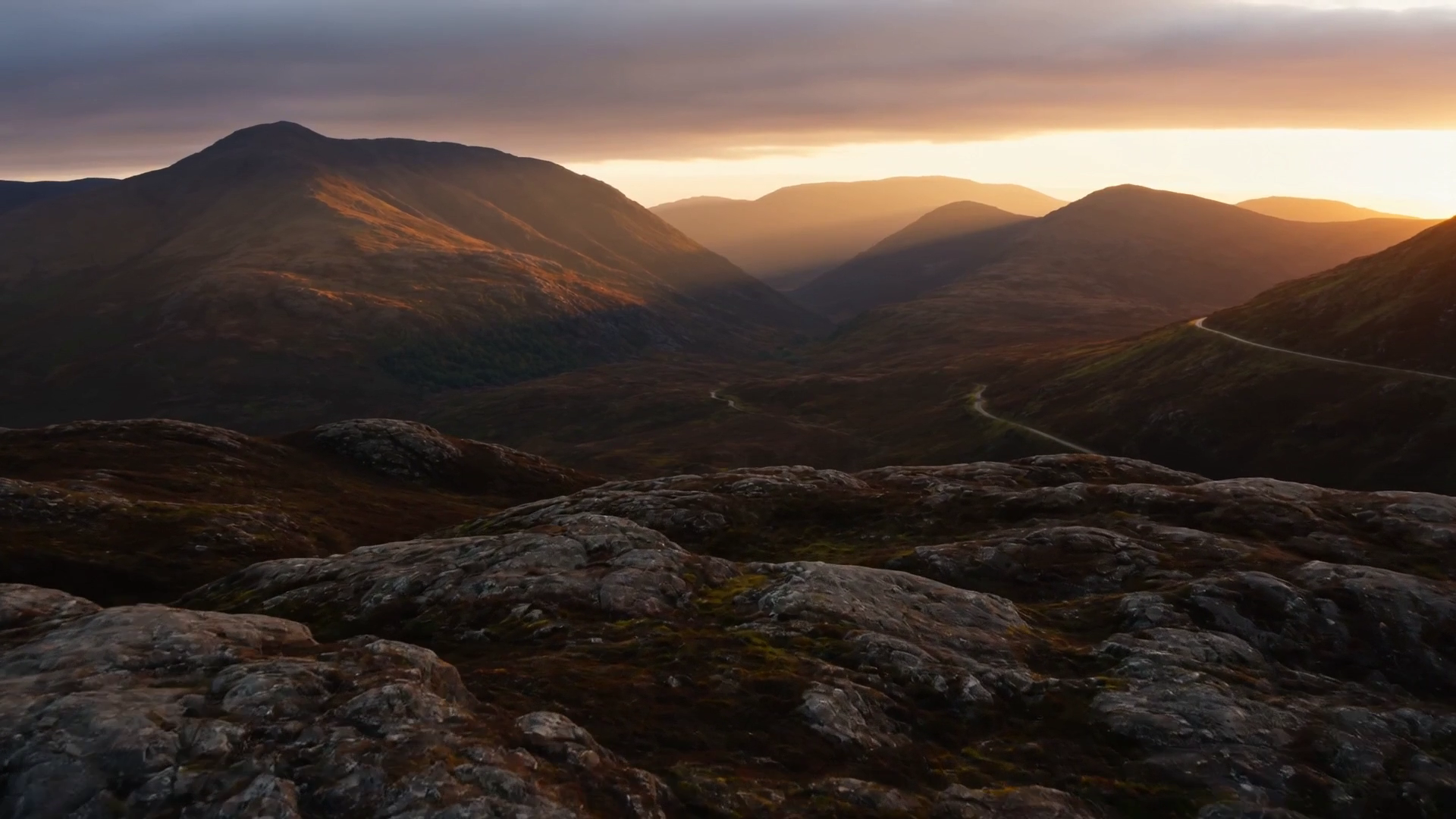 Aerial view of a Highland glen at golden hour, a single-track road winding through the valley toward distant mountains.