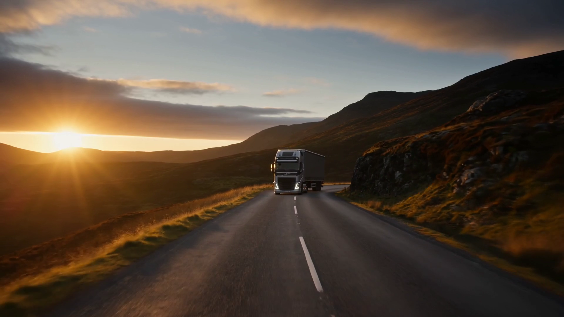 A silver articulated truck travelling along a single-track Highland road, low sun flaring over the hills to the left.