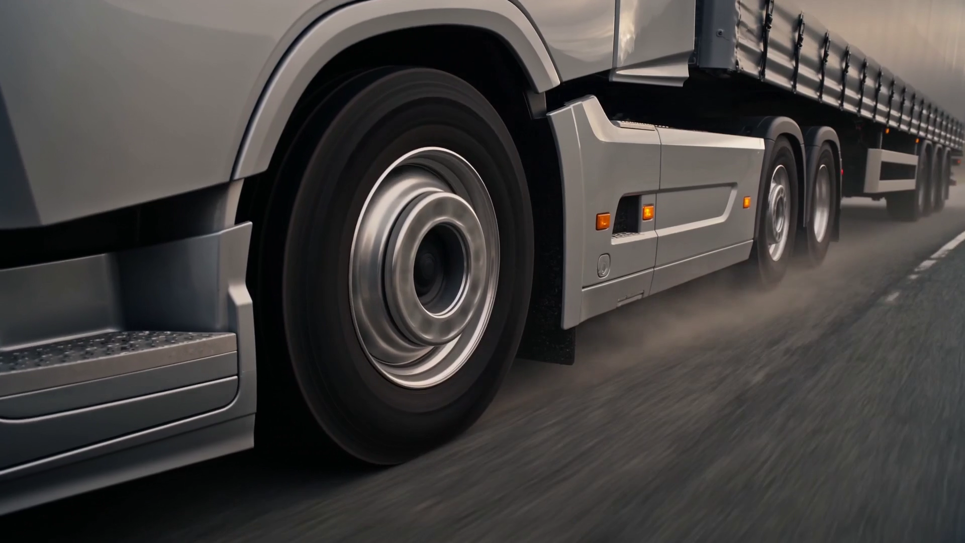 Low-angle view of a truck's spinning drive wheel and curtain-side trailer, the road blurring with speed.